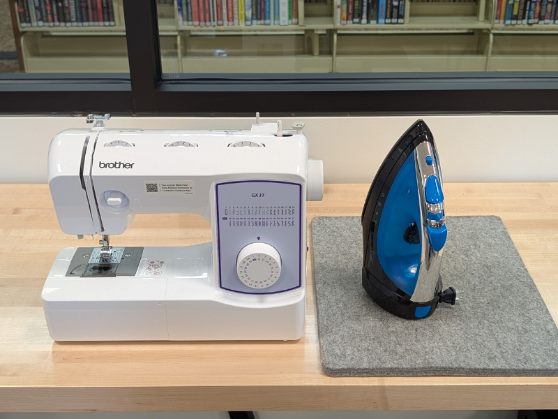 A white sewing machine and a blue and black iron sitting upright on a wooden table with a bookcase in the background.