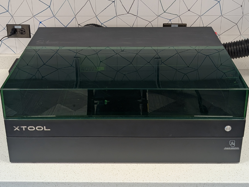 A black XTool S1 Laser Engraver machine sitting on a white countertop with white and grey backsplash behind the machine.
