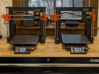 Two Prusa MK4S+ 3D Printer machines sitting on a wooden desk with a window and blinds in the background